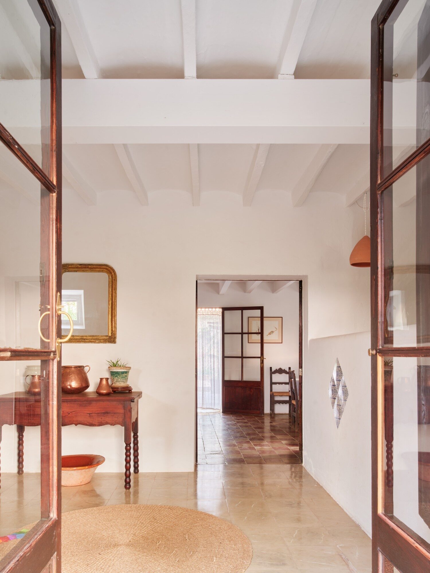 The entrance to the main house: glass doors open onto a hall with beamed ceiling, copper jugs on a wooden table and a doorway beyond